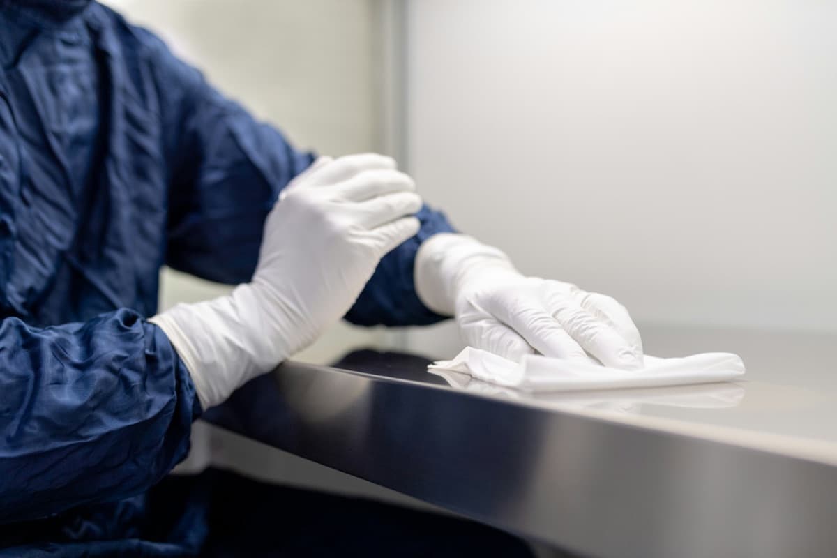 Technician in a gown and gloves wiping down a stainless steel cleanroom surface